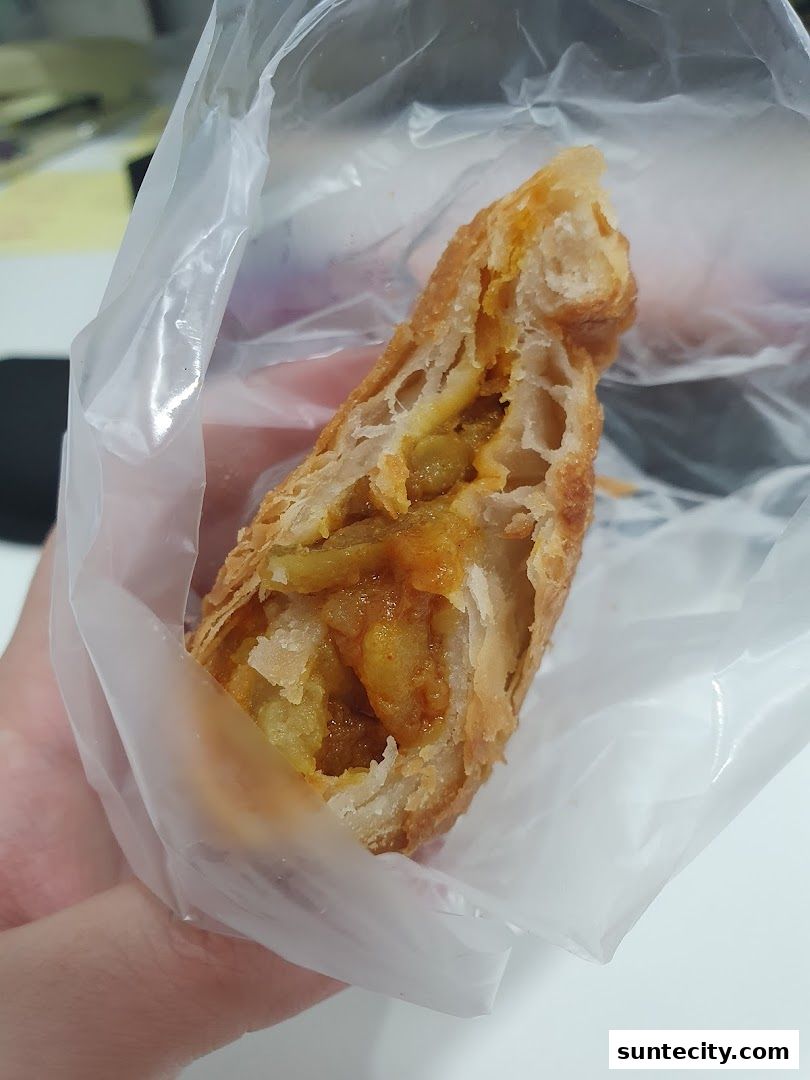 A hand holds a savory pastry filled with spiced potatoes and peas, wrapped in plastic.