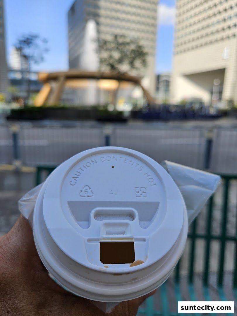 A person holds a takeaway coffee cup with a lid, with a blurred urban background.