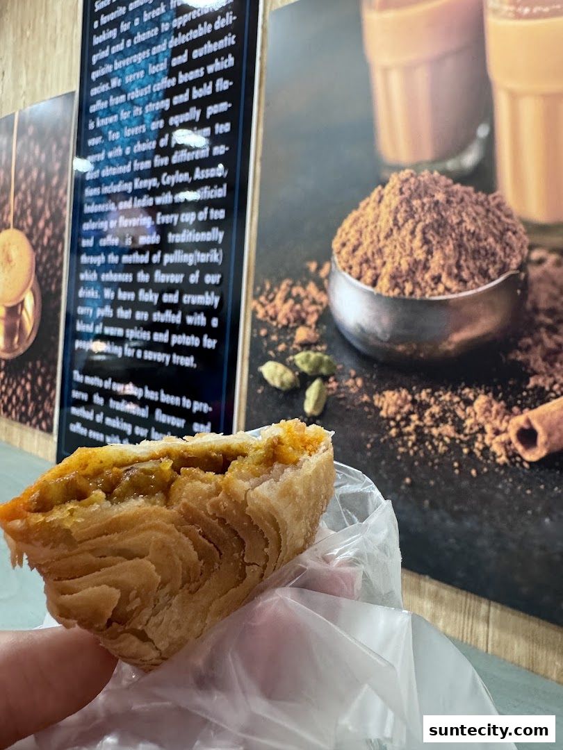 A close-up of a flaky curry puff being held, with a background showcasing tea and coffee ingredients.