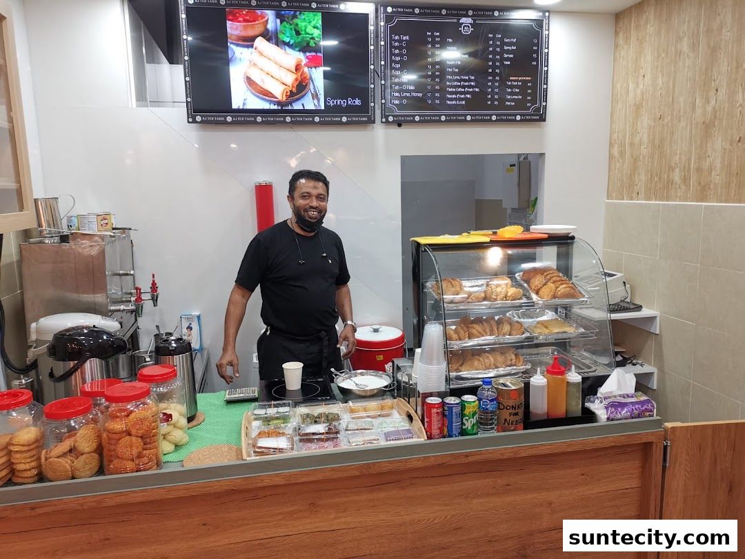 A smiling man stands behind a counter at AJs Teh Tarik, with a display of pastries and drinks.