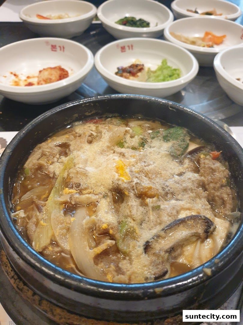 A steaming bowl of Korean stew with various side dishes in the background.