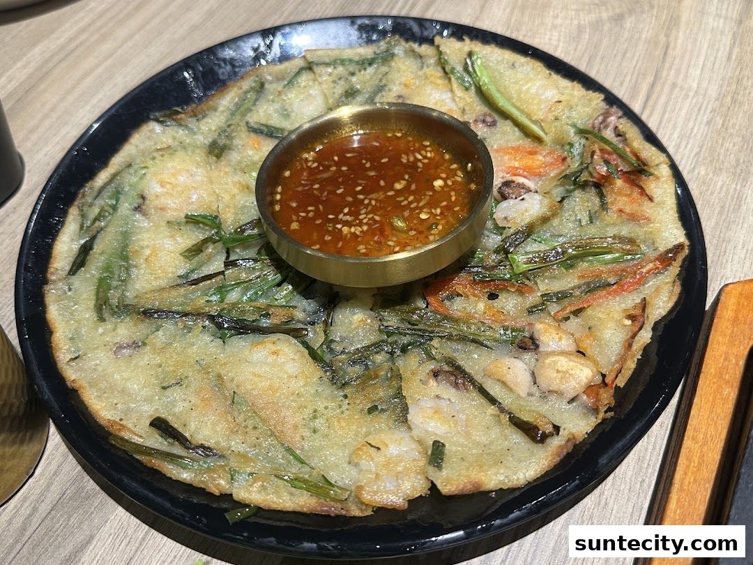 A close-up of a savory Korean pancake with seafood and vegetables, served with dipping sauce.