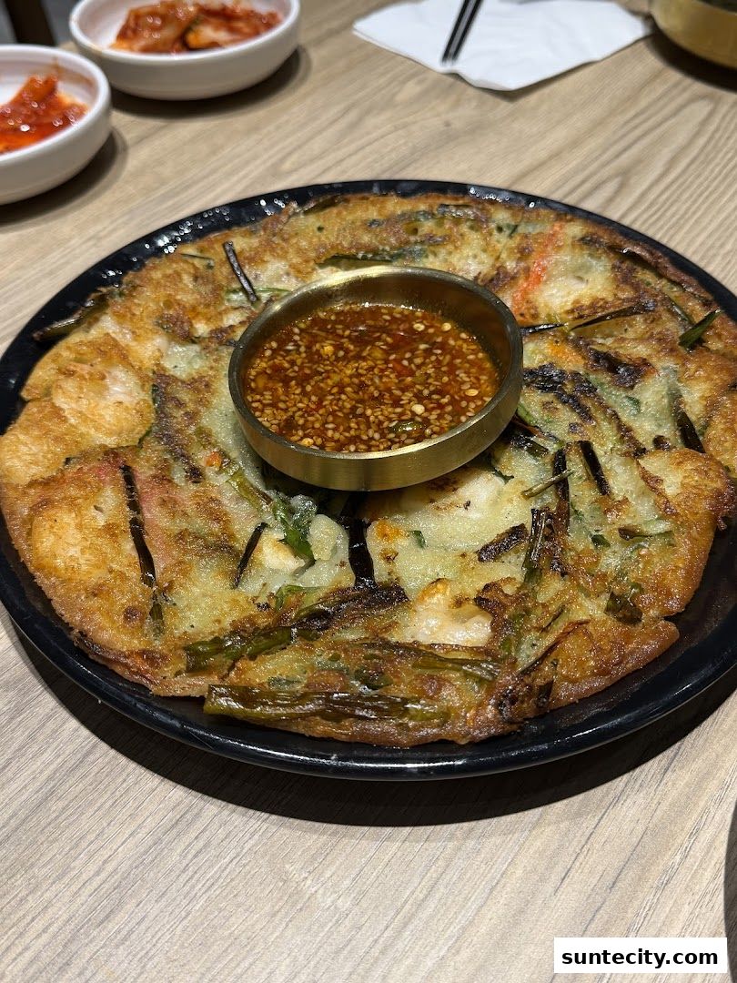 A close-up of a savory Korean pancake with dipping sauce and side dishes.