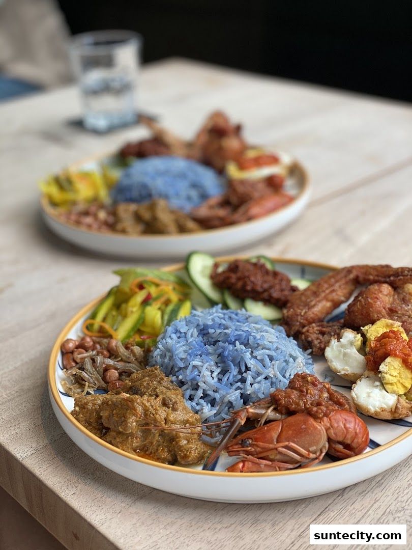 Two plates of nasi kerabu with blue rice, fried chicken, and various side dishes.