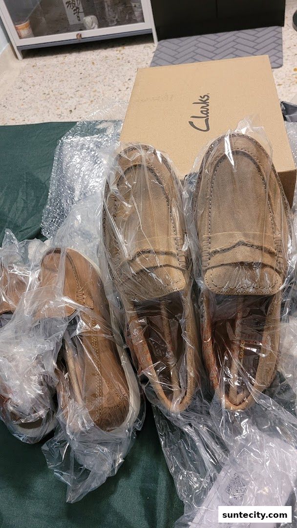 Several pairs of brown suede loafers are displayed, some in plastic packaging, with a Clarks shoebox in the background.