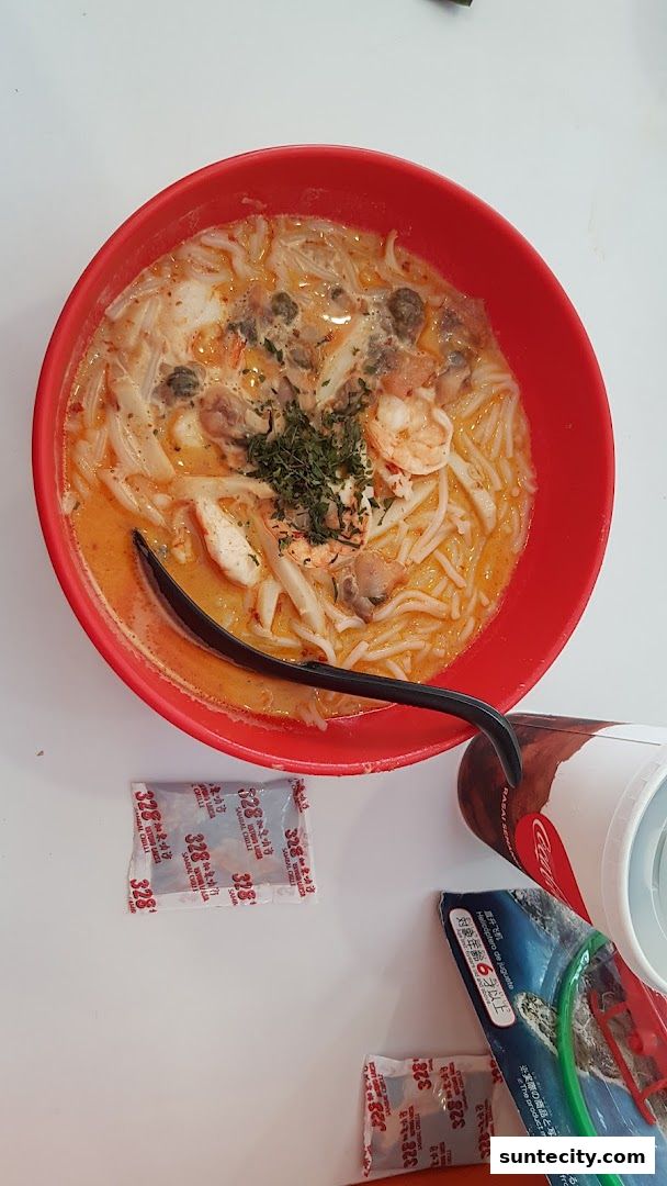 A bowl of laksa with shrimp, clams, and herbs, served with chili packets and a drink.