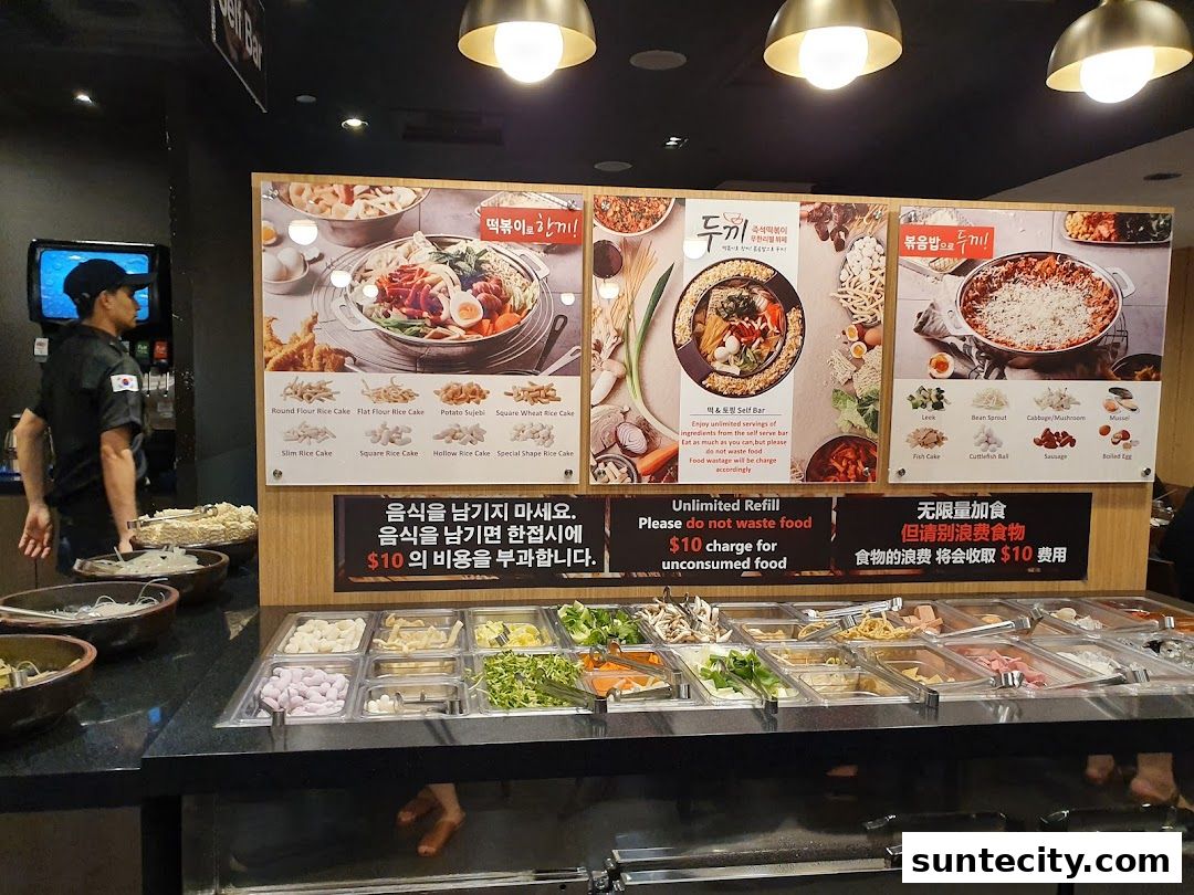 A display of ingredients for Korean tteokbokki with a sign about unlimited refills and no waste.