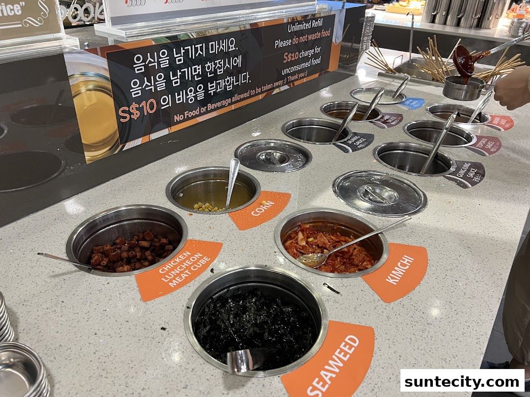 A food bar with various ingredients like chicken, corn, kimchi, and seaweed, with sauces being served.