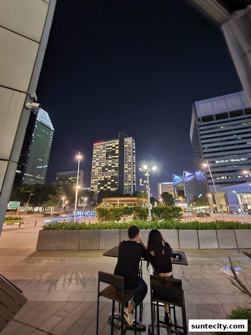 Couple sitting at a table outside a building at night with city lights in the background.