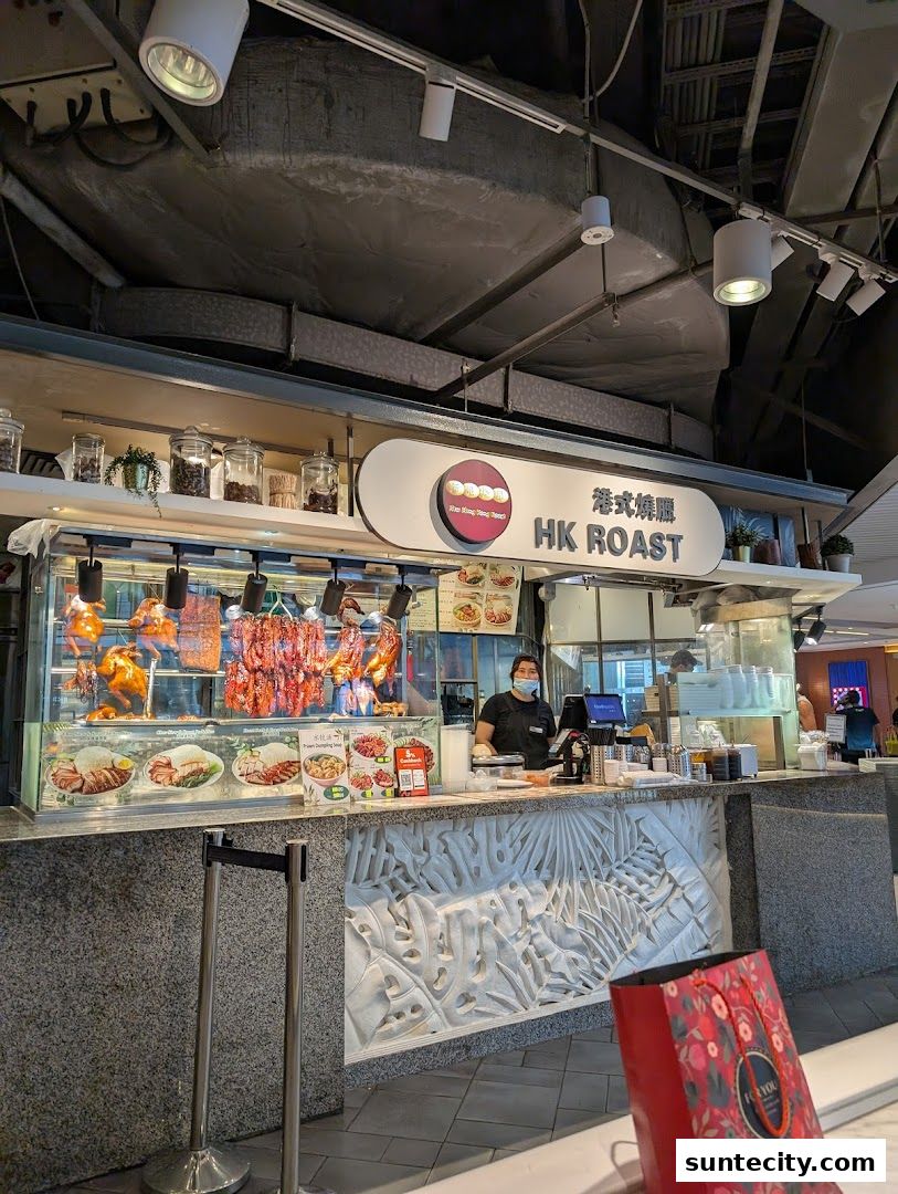 A food stall with roasted meats displayed, a staff member behind the counter, and a shopping bag in the foreground.