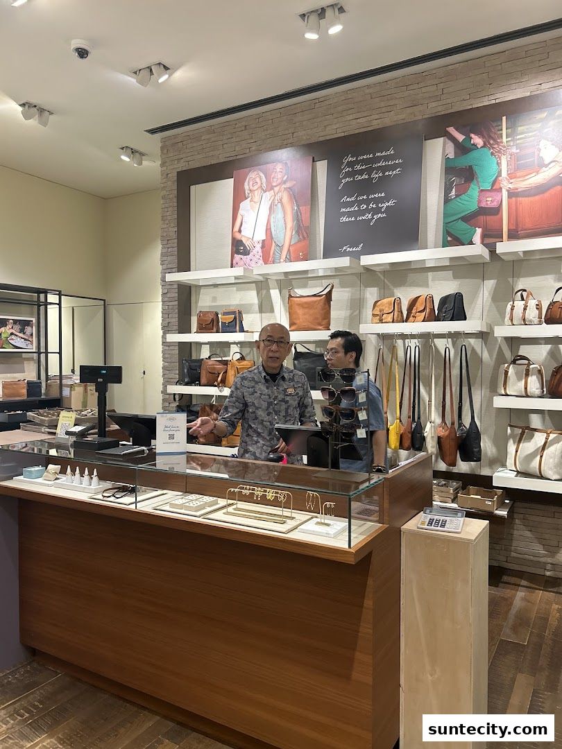 Two staff members stand behind a counter in a Fossil store, with bags displayed on shelves.
