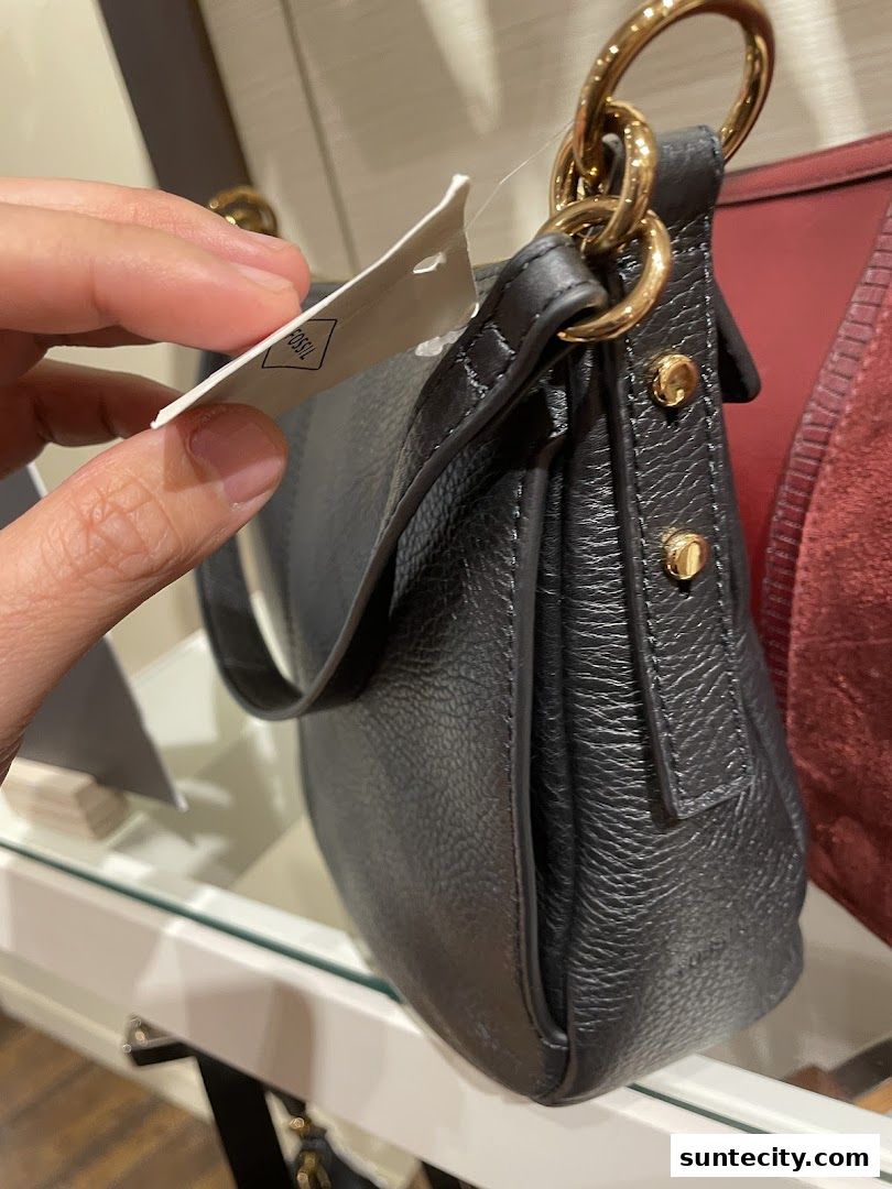 A hand holds a tag on a black leather Fossil handbag with gold hardware.