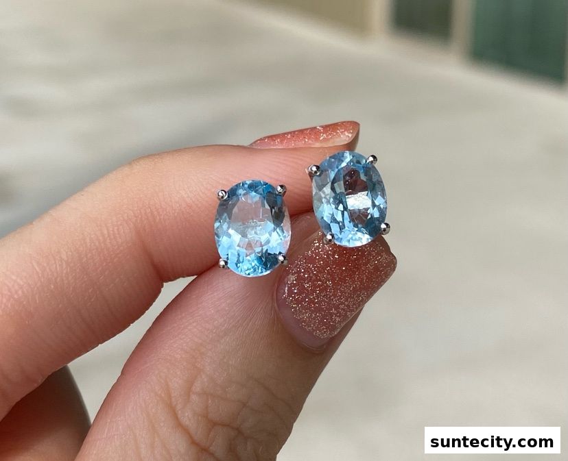 A close-up shot of a pair of oval-cut blue gemstone stud earrings held by a hand.