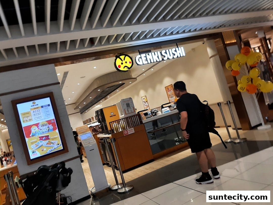 A man stands outside Genki Sushi Suntec City, with a digital menu board and balloons.
