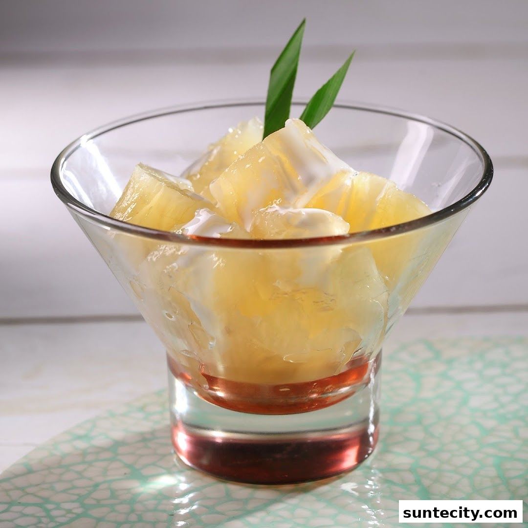 A close-up of a dessert in a glass, featuring chopped fruit with a creamy sauce and pandan leaves.