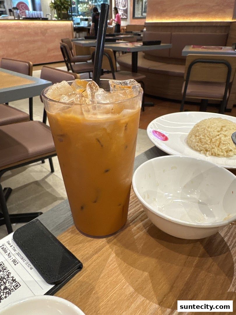A tall glass of Thai iced tea with ice sits on a wooden table next to a plate of rice.