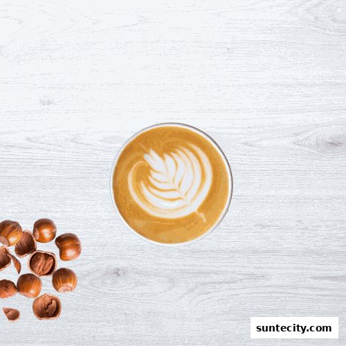 A top-down view of a latte with intricate foam art next to a pile of hazelnuts.