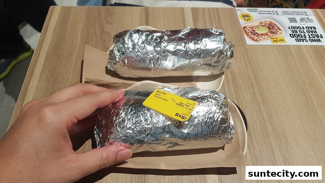 Two foil-wrapped burritos are presented on a wooden table, with a hand reaching for one.