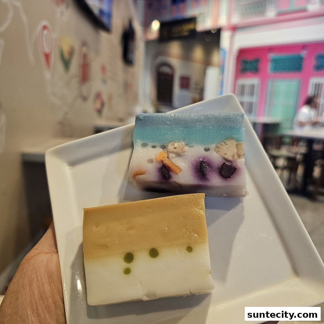 Two colorful, layered desserts are presented on a white plate, with a blurred background of a vibrant shop interior.