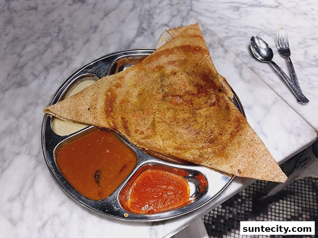 A triangular dosa served with three dipping sauces on a metal plate with cutlery.