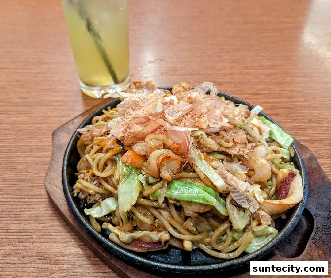 A sizzling plate of yakisoba with shrimp, squid, and cabbage, topped with bonito flakes, served with a drink.