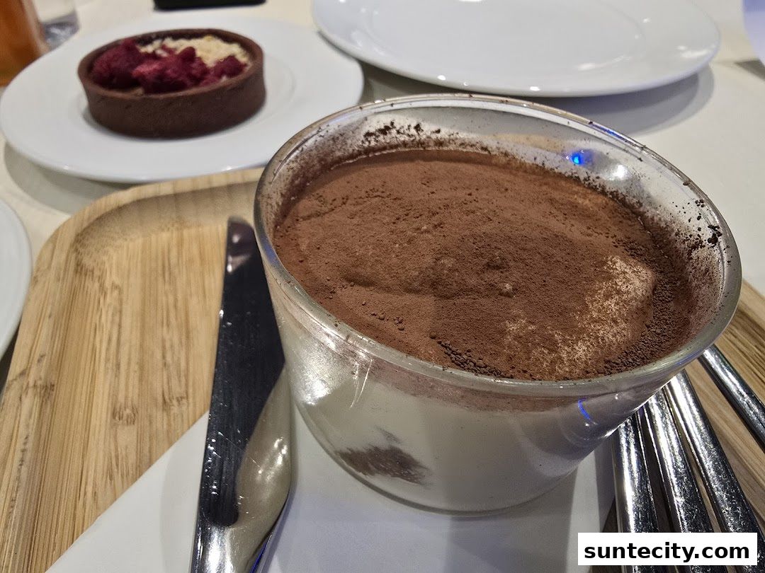 A close-up of a dessert in a glass, dusted with cocoa powder, with a tart in the background.