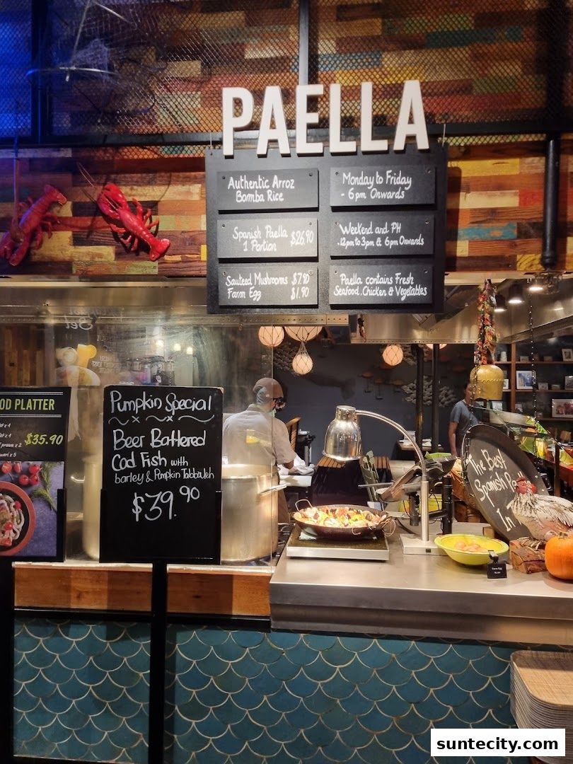 A food stall counter with a 'PAELLA' sign, menu boards, and a chef preparing food.