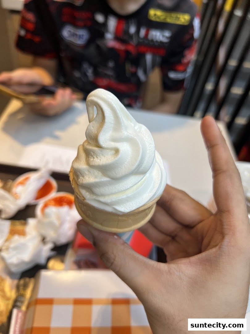 A hand holds a soft-serve ice cream cone with a person in the background.