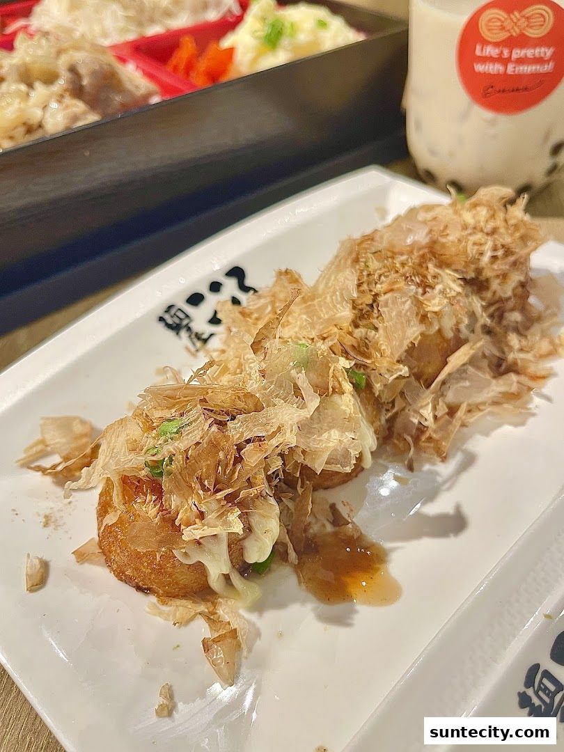 A close-up of takoyaki topped with bonito flakes and sauce, with a bento box and drink in the background.