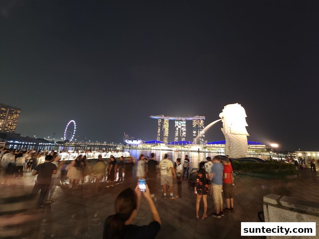 A crowd of people gather at night to view the illuminated Merlion statue and city skyline.