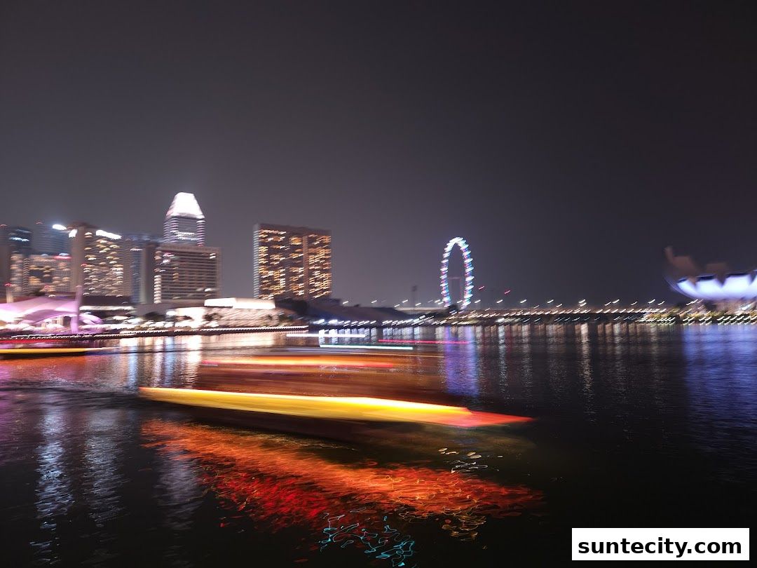 Night view of a city skyline with a river and boats, featuring illuminated buildings and a Ferris wheel.