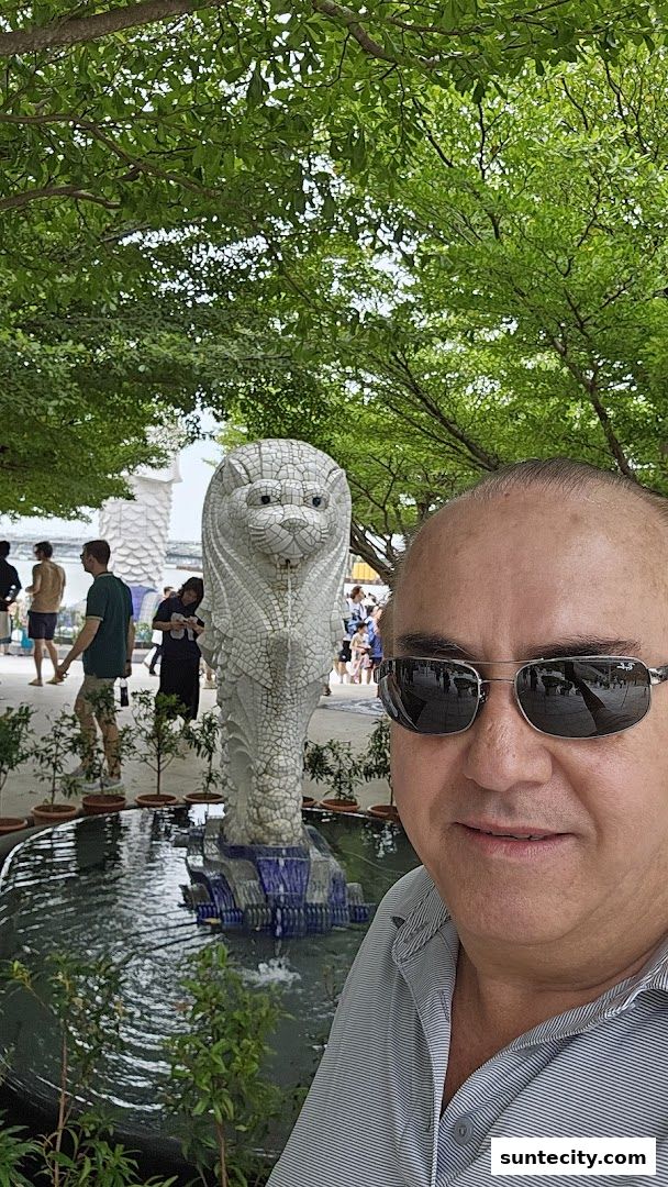 A man takes a selfie in front of a Merlion statue and fountain.