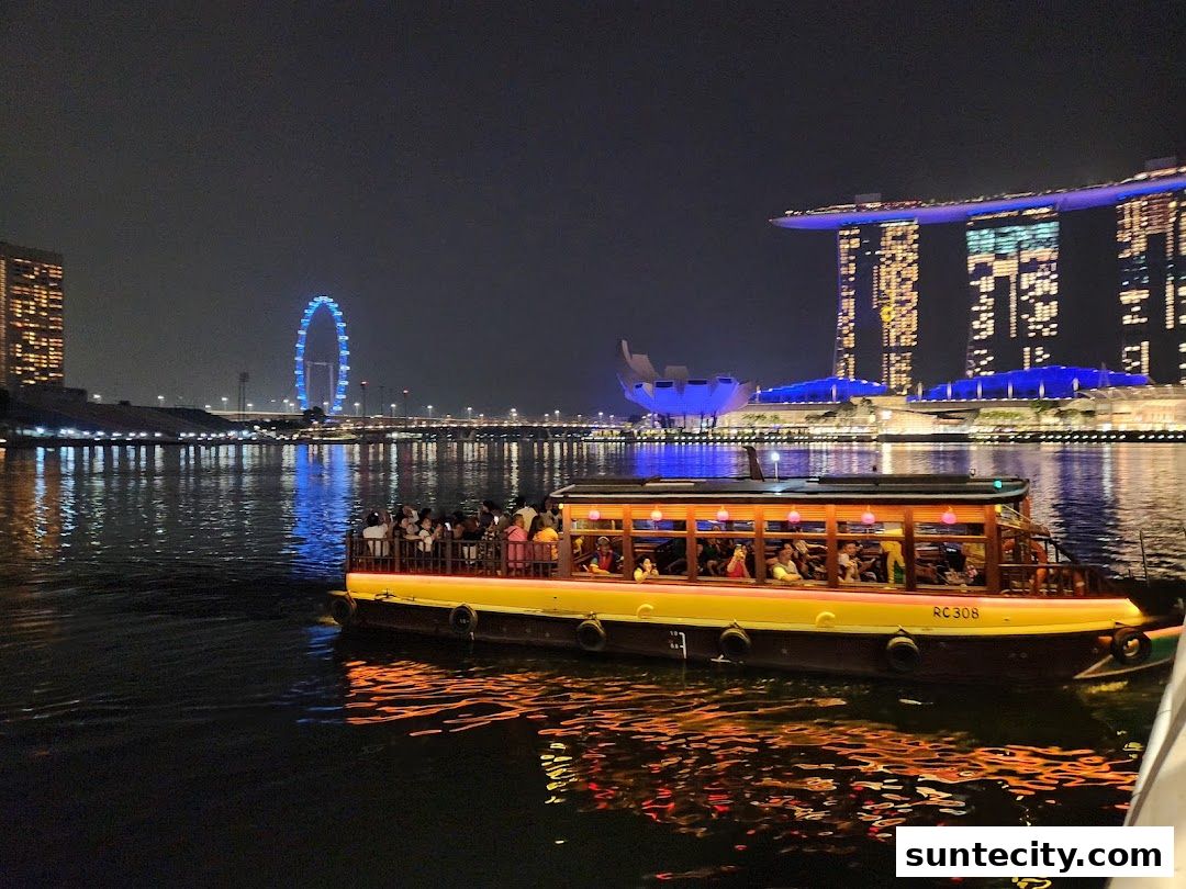 A boat filled with people cruises on the water at night with city skyline in the background.