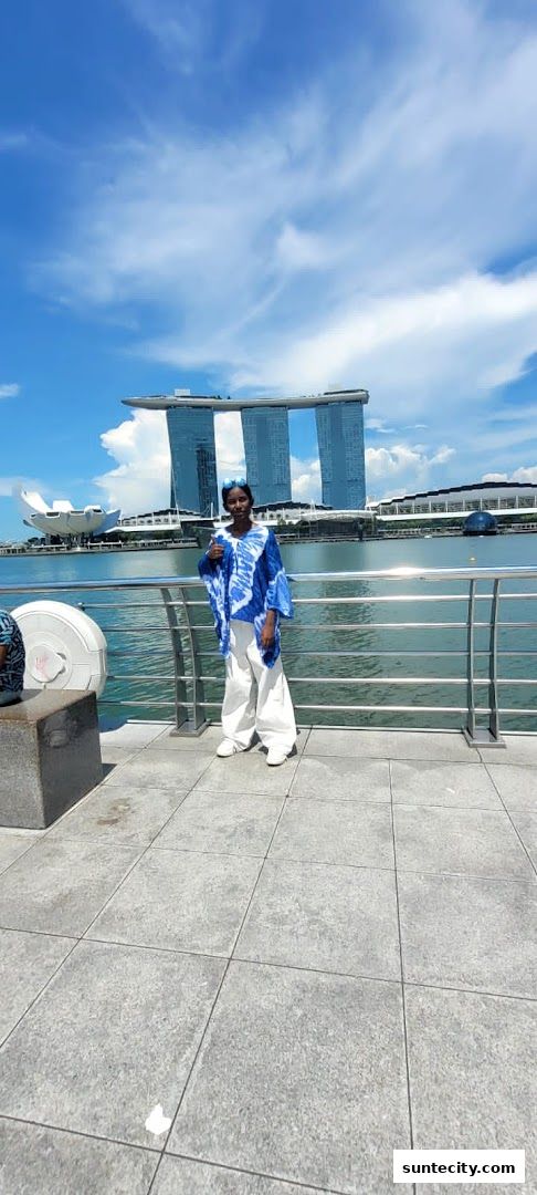 A person in a blue and white outfit stands by a railing with a famous Singapore landmark in the background.