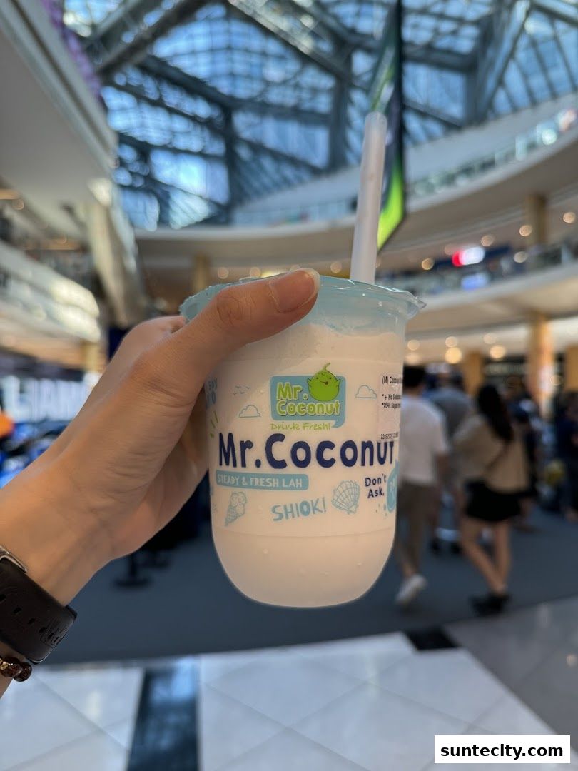 A hand holds a Mr. Coconut drink with the mall interior blurred in the background.