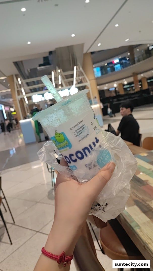 A hand holds a Mr. Coconut shake with a straw in a shopping mall.