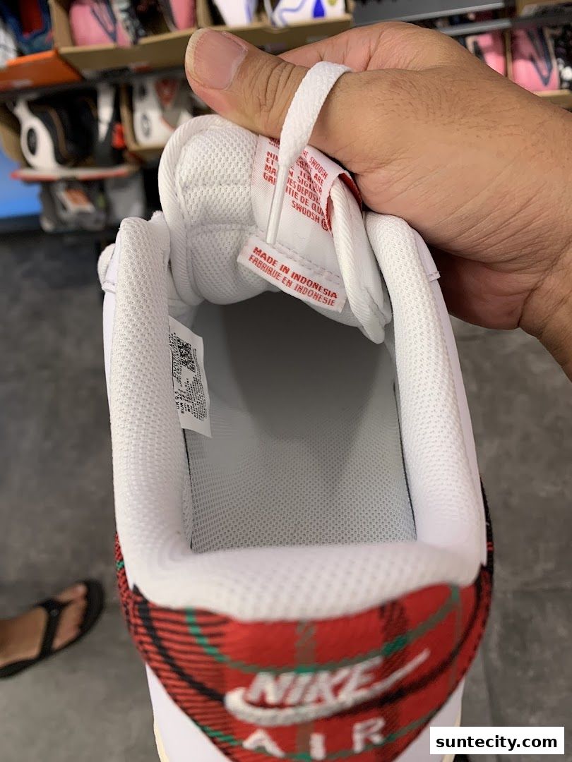A hand holds a white Nike Air sneaker with a red plaid heel tab.