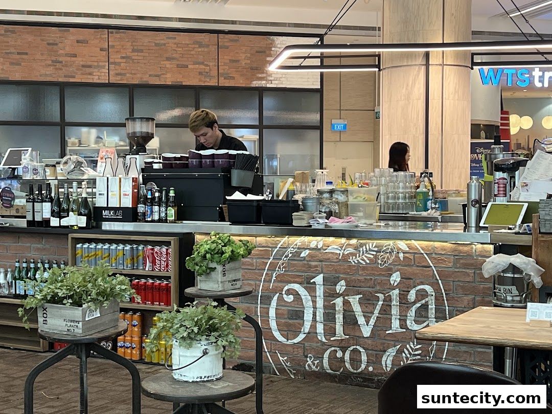 A barista works behind the counter at Olivia & Co., a cafe with brick accents and drinks on display.