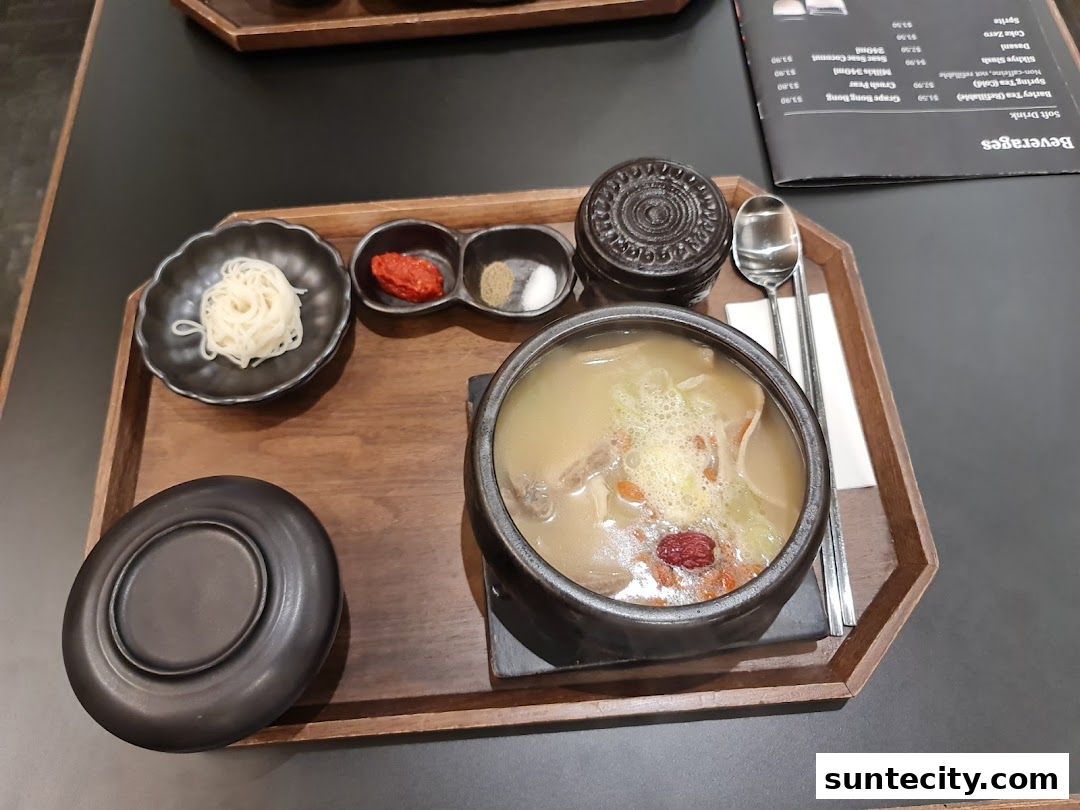 A bowl of Korean soup with noodles, condiments, and a beverage menu on a tray.