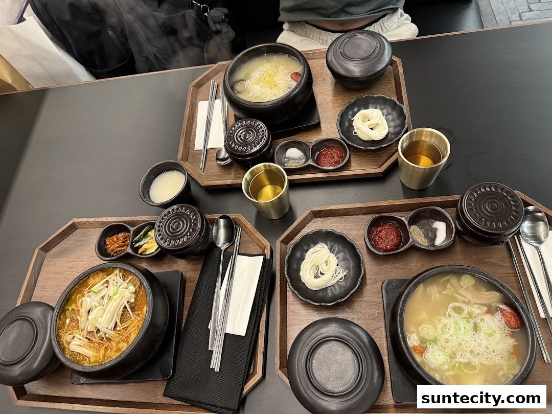 Two steaming bowls of Korean soup with side dishes and noodles on wooden trays.