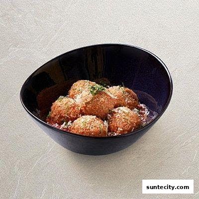 A dark bowl filled with golden-brown meatballs in tomato sauce, topped with cheese and herbs.