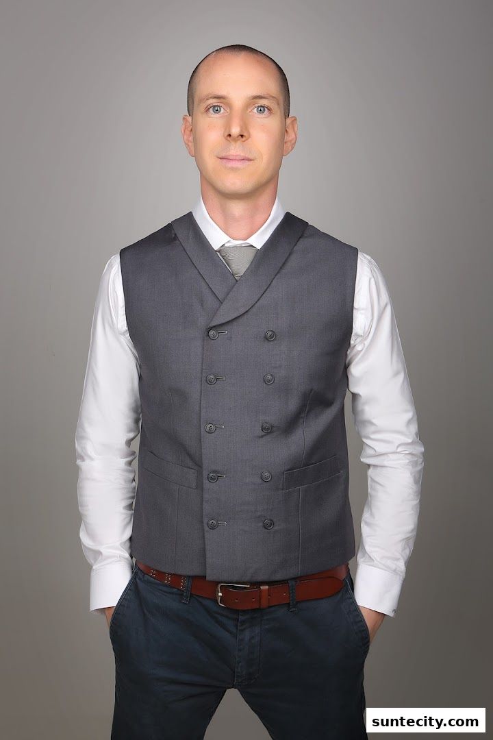 A man in a grey double-breasted vest, white shirt, and blue pants stands with hands in pockets.
