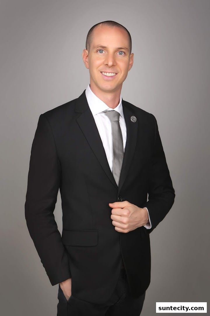 A man in a black suit and tie smiles against a gray background.