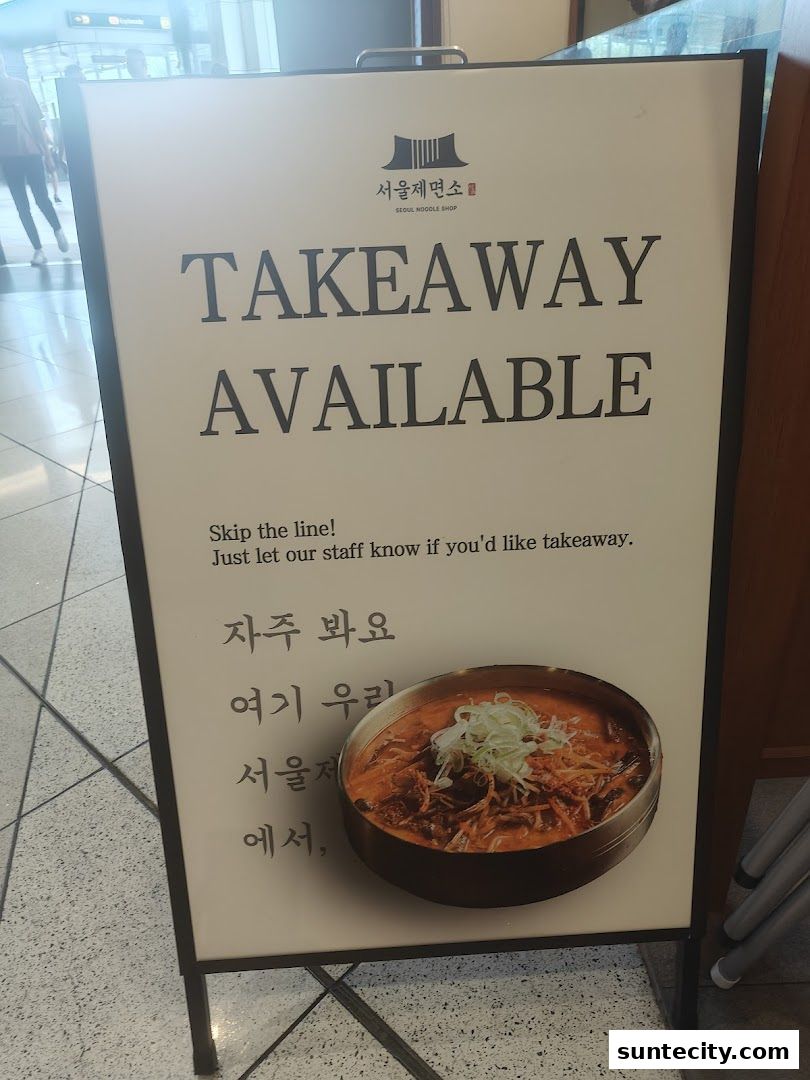 A sign outside Seoul Noodle Shop advertising takeaway availability with a picture of a noodle dish.