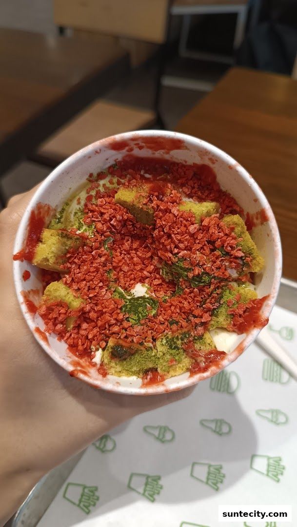 A hand holds a cup of frozen custard topped with cake pieces, strawberry sauce, and red sprinkles.