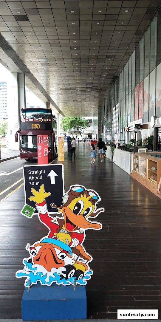 A cartoon duck mascot stands on a wooden walkway next to a double-decker bus.
