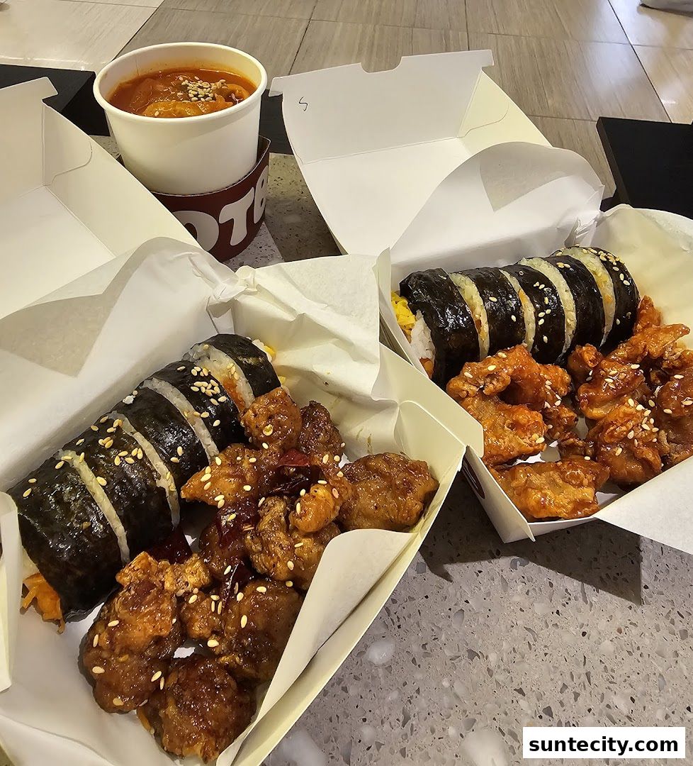 Two boxes of Korean food, featuring kimbap and fried chicken, with a cup of soup.