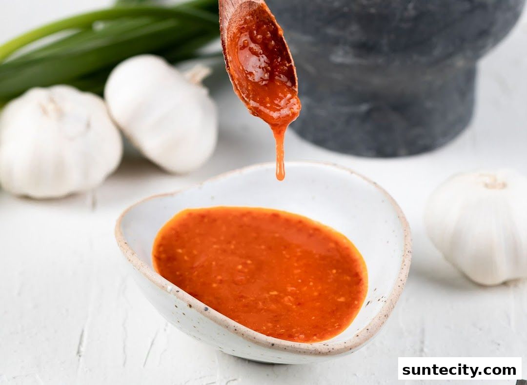 A spoon drips a vibrant red sauce into a bowl, with garlic and green onions in the background.