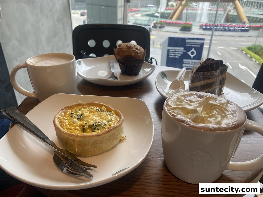 A table with Starbucks coffee, quiche, muffin, and chocolate cake with a city view.
