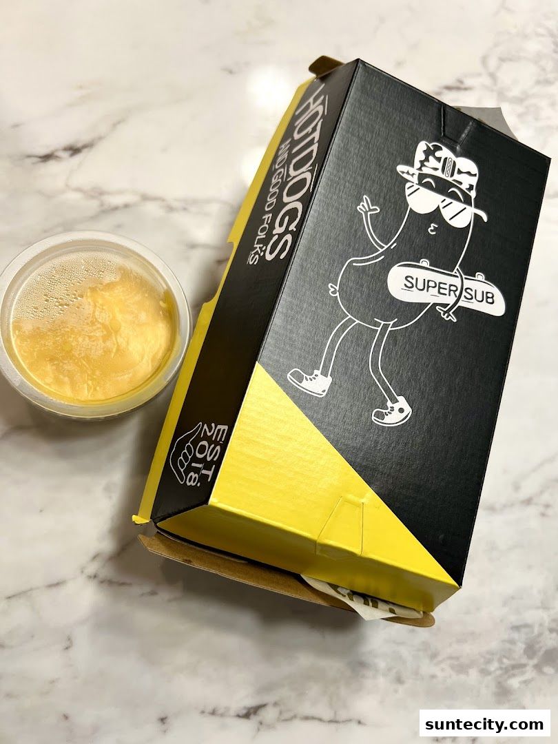 A black and yellow Super Sub hotdog box with a cartoon character and a side of sauce.